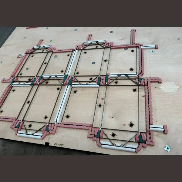 Wooden die forme board featuring a grid of pink and white sections reinforced with black truss strings, white rods, and green inserts, marked with spider-like drill symbols and labeled “23 GRIP” at the base