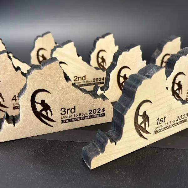 Wooden trophies shaped like stylized waves or mountains, engraved with surfer silhouettes and event details for Lion Rock Boardriders youth surf competitions.